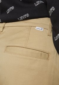 Pantalons en coton beige avec une poche arrière à couture horizontale, une étiquette Levi's, et un haut noir avec des logos Levi's blancs répétés.