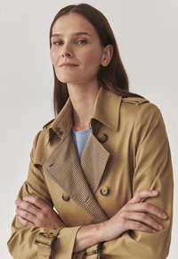 Beige trench coat with a houndstooth collar, double-breasted buttons, and belt buckle accents, featuring a smooth texture and tailored fit.