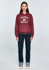 Sudadera burdeos con "BIG STAR DNM CLUB" impreso en blanco, con un bolsillo frontal y puños acanalados, combinada con jeans de mezclilla oscura.