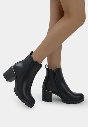 Bottines noires à talon épais, en matière synthétique lisse, avec fermeture éclair sur le côté et accents métalliques subtils. Semelle profilée pour une meilleure adhérence.
