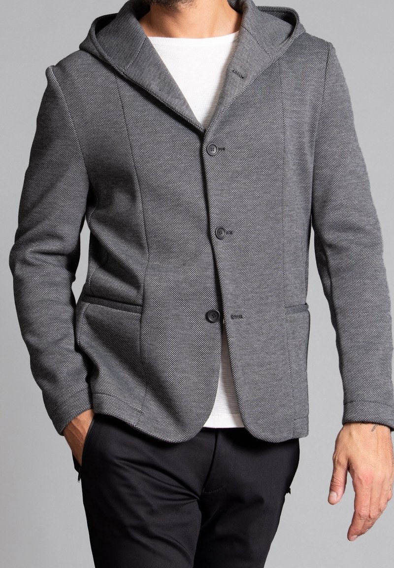 Blazer grigio in maglia con cappuccio, tessuto testurizzato, chiusura a due bottoni, tasche laterali e vestibilità slim, abbinato a una camicia bianca sotto.