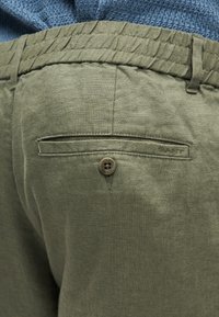 Παντελόνι από λινό σε ελιά πράσινο με ελαστική μέση και πίσω τσέπη με κουμπί, φορεμένο με μπλε πουλόβερ με υφή.