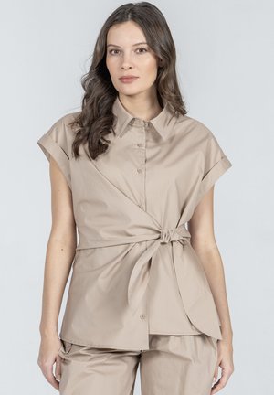 Kobieta z długimi falowanymi włosami nosi beżową bluzkę na krótki rękaw z guzikami, związaną w talii, oraz pasujące beżowe spodnie na jasnym tle.