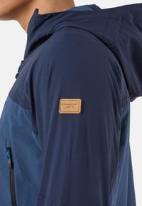 Man draagt een marineblauwe capuchonjas met een Swedemount-logo op de bovenarm en een met rits afgesloten zak op de borst.