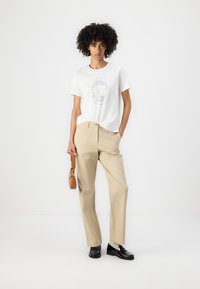 Hvid T-shirt med grafisk print, beige lige ben bukser og sorte loafers. Modellen holder en lille brun håndtaske. Simpelt, afslappet outfit.