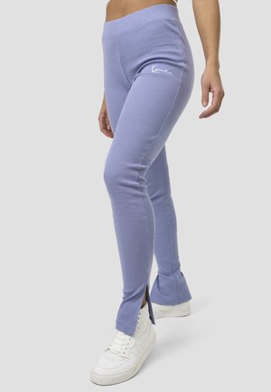 Frau trägt lavendelfarbene, gerippte Leggings mit seitlichen Schlitzen und weißen Sneakers, steht vor einem schlichten hellgrauen Hintergrund.