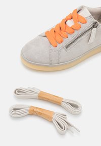 Basket en suede gris clair avec des lacets orange et une fermeture éclair sur le côté, accompagnée de deux remplacements de lacets blancs fixés avec du ruban adhésif.