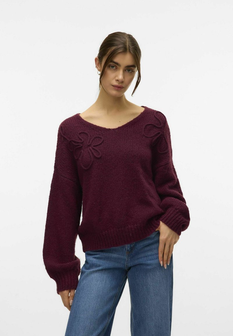 Bordeaux gebreid sweater met een V-hals, voorzien van bloemenborduurwerk op de schouders en geribbelde manchetten en zoom. Gecombineerd met blauwe wijde jeans.