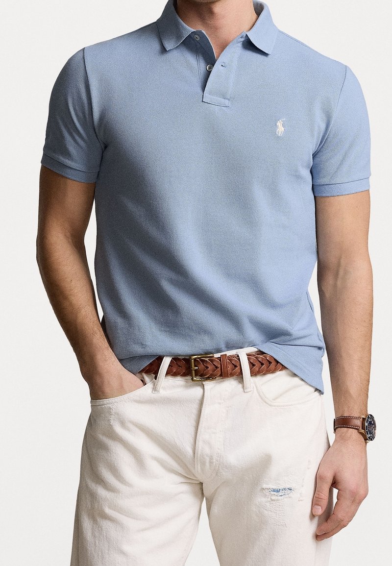 Homme portant un polo bleu clair à manches courtes, ceinture tressée marron, pantalon blanc avec une petite déchirure et montre-bracelet marron, main dans la poche.