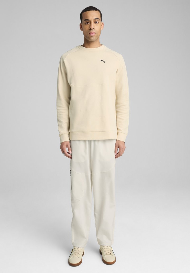 Sudadera beige con cuello redondo y logotipo negro, combinada con pantalones crema que tienen bolsillos laterales. Los zapatos son ligeros con una suela marrón.