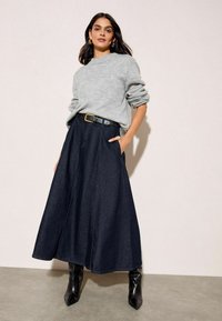 Sötétkék farmer szoknya magas derékkal, széles pliszével és zsebekkel, világosszürke pulóverrel és fekete magassarkú csizmával párosítva.