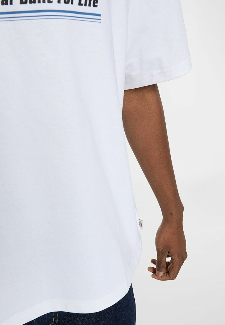 Dark-skinned arm hanging beside a white short-sleeve shirt with partial black and blue text, paired with dark blue jeans.
