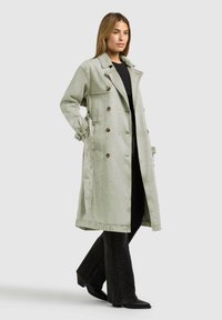 Nő világoszöld hosszú ballonkabátban, fekete felsőben, bőszárú fekete nadrágban és fekete hegyes orrú csizmában, egyik kezét a zsebében tartva áll.