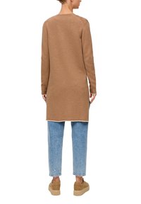Un long pull en tricot marron avec un col rond, des poignets côtelés et des fentes sur les côtés, assorti à un jean en denim bleu clair et des baskets beige.