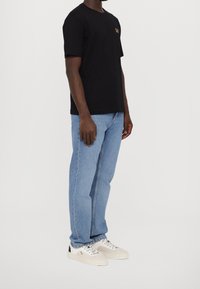 Camiseta de algodón negra con un logo en el pecho, combinada con unos vaqueros de denim azul claro y zapatillas beige con detalles en negro.
