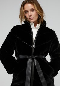 Giacca in pelliccia sintetica nera con colletto alto, chiusura con zip e motivo a zig-zag. Include una cintura nera in pelle rimovibile per la regolazione.