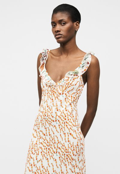 Γυναίκα με κοντά μαλλιά, φορέμα αμάνικη λευκή με πορτοκαλί ανθικό σχέδιο και λαιμόκοψη με βολάν, που στέκεται μπροστά σε απλό φόντο.