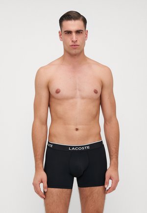 Boxer brevissimi neri in materiale elastico con un cinturino bianco marchiato "LACOSTE." Texture liscia, design aderente, lunghezza media.