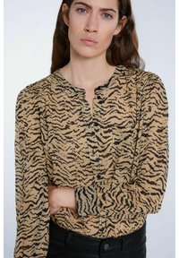 Beige Bluse mit schwarzem Tigerstreifenmuster, runder Ausschnitt, Knopfverschluss, langen Ärmeln und einer Brusttasche. Kombiniert mit schwarzen Hosen.