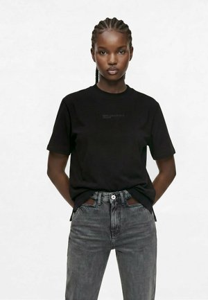 Femme aux cheveux tressés portant un t-shirt noir à manches courtes et un jean gris délavé, debout les mains dans le dos.