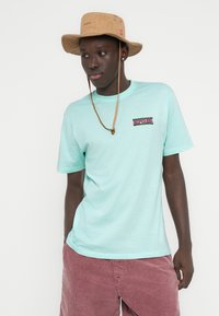 Ανοιχτό τιρκουάζ κοντομάνικο μπλουζάκι με πολύχρωμο λογότυπο "Ripcurl". Συνδυασμένο με ροζ βερμούδα από κορδόνι και μπεζ καπέλο τύπου bucket με κορδόνι στο πηγούνι.