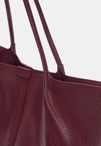 Burgundy nahka-tote-laukku, jossa on teksturoitu pinta, kaksi pitkää kahvaa ja avoin yläosa, hienovaraisilla ompeleilla ja ilman näkyvää metalliosaa.