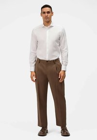 Camicia bianca a maniche lunghe con colletto button-down, abbinata a pantaloni marroni plissettati. Il tessuto appare liscio con una vestibilità sartoriale.