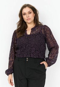 Lilla bluse i blonder med lange, puffede ærmer og en nøglehul-udskæring. Båret med sorte bukser med striber og to knapper.