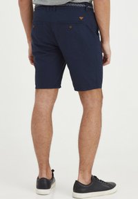 Navy blue shorts with a textured fabric, featuring two back pockets with buttons and a triangle logo detail. Paired with black sneakers.