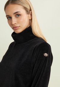 Suéter de cuello alto de pana negra con tejido texturizado, forma ajustada y un detalle de logo en oro rosado en la manga izquierda.