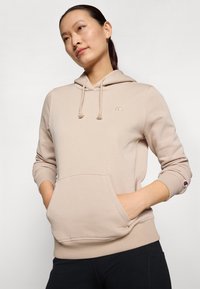 Hoodie beige en tissu doux avec une poche avant, capuche à cordon de serrage et détail de logo sur la poitrine et la manche. Présente des poignets et un ourlet côtelés.