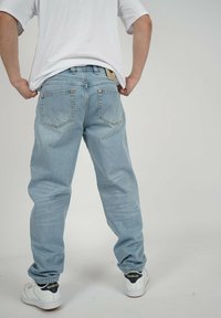 Person in hellblauen Jeans und weißen Reebok-Sneakers, die ein weißes T-Shirt leicht nach unten ziehen, stehend vor einem schlichten hellen Hintergrund.