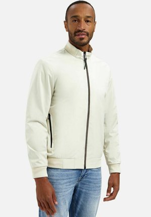 Man draagt een lichtbeige jas met rits, geribbelde boorden en kraag, gecombineerd met blauwe spijkerbroek, staand tegen een effen achtergrond.