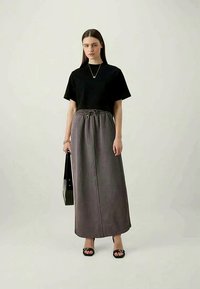 Femme avec de longs cheveux raides portant un haut noir à manches courtes, une jupe maxi grise à cordon, des sandales noires, portant un sac à main rectangulaire noir.