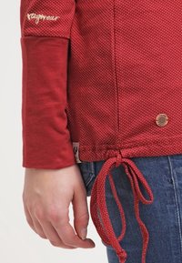 Röd mönstrad långärmad topp med dragsko i midjan, med en logotyp på ärmen och en etikett vid fållen, tillverkad av mjukt tyg.