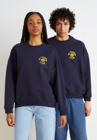 Ναυθένιοι μπλούζες με  ribbed μανσέτες και στρίφωμα, περιλαμβάνοντας κίτρινη κεντητή επιγραφή "On Vacation Club". Το μοντέλο είναι ντυμένο με ανοιχτό μπλε τζιν.