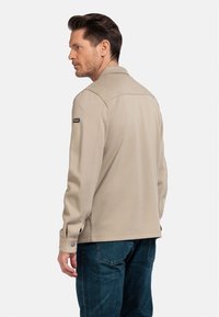 Beige jas met een kraagontwerp, lange mouwen en knoopmanchetten; gecombineerd met blauwe denim jeans. De textuur lijkt glad.