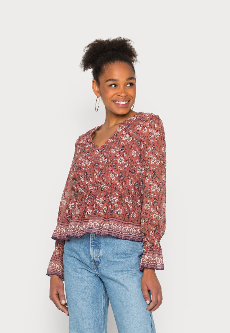 Blusa de manga longa em terracota com decote em V e estampa floral; tem uma cintura elástica e um bordado decorativo na bainha, além de punhos amplos. Combinada com jeans azul claro.