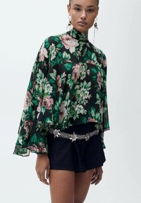 Blommig blus i svart med gröna och rosa rosor, hög hals, flödande ärmar, bärs över mörka shorts, kompletterad med ett dekorativt kedjebälte.