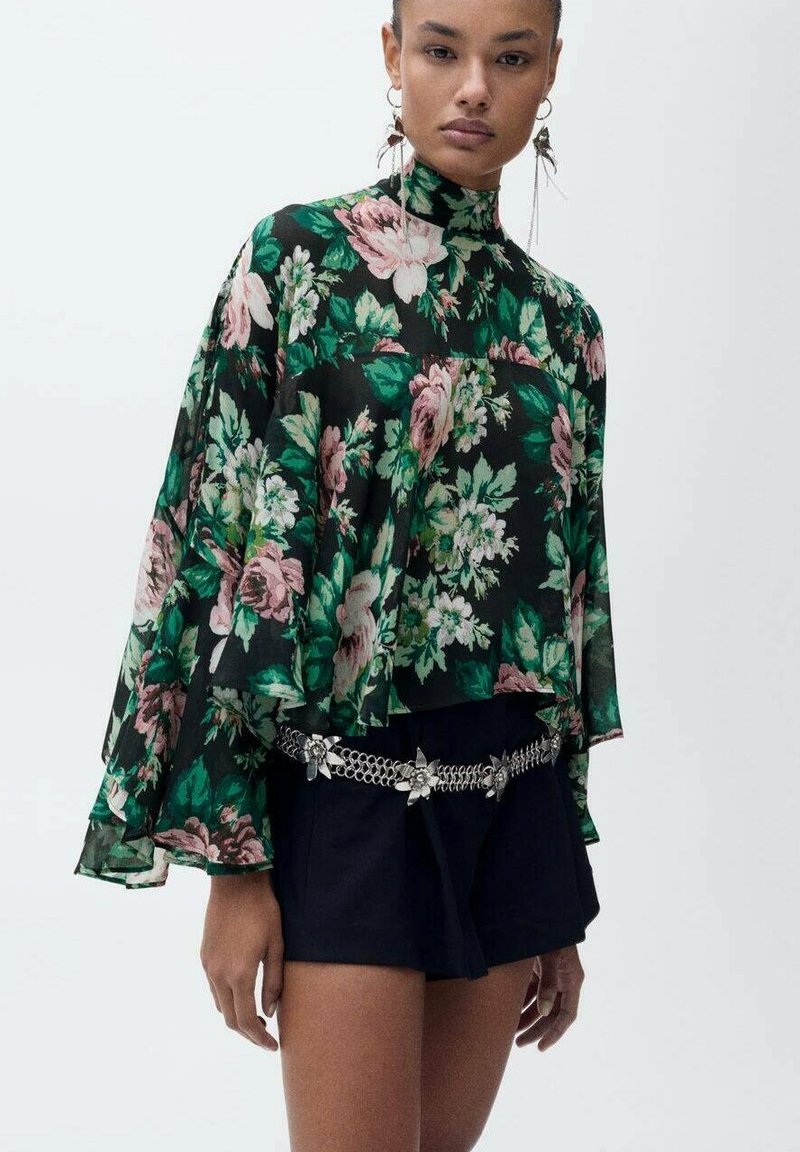 Blommig blus i svart med gröna och rosa rosor, hög hals, flödande ärmar, bärs över mörka shorts, kompletterad med ett dekorativt kedjebälte.