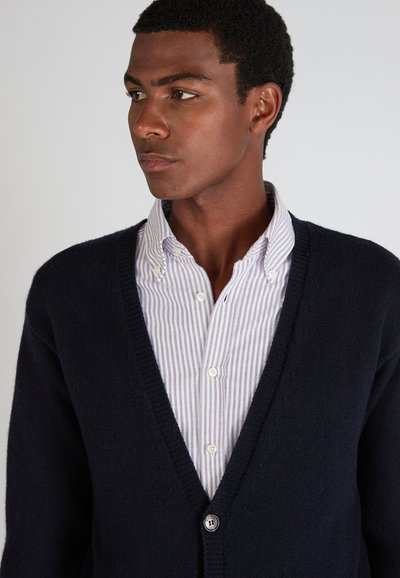 Cardigan en laine bleu marine avec un col en V, doté d'une fermeture à boutons, porté sur une chemise à col rayée violet clair et blanche.