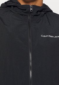 Chaqueta negra con capucha y cremallera frontal, hecha de material ligero. Presenta un logo de la marca en blanco en el área del pecho izquierdo.