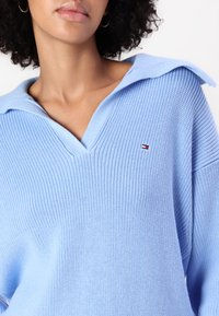 Pulover light blue cu tricot texturat, având decolteu în V și guler larg. Include un detaliu mic cu logo pe piept. Material moale.