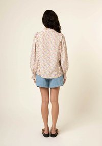 Hauchdünne Bluse mit dezentem Muster aus weichem Stoff und Rüschenschultern, kombiniert mit Denim-Shorts. Das Model steht mit dem Rücken zur Kamera und zeigt einen lässigen Look.