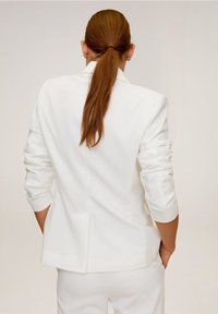 Blazer blanco de corte entallado, con mangas largas y abertura en la parte trasera, combinado con pantalones a juego. Textura de tela lisa.