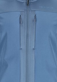 Blå zip-up jakke med glat stof, ribbet øverste panel, dobbelt snoretræk og et logo på brystet. Funktionelt og enkelt design.