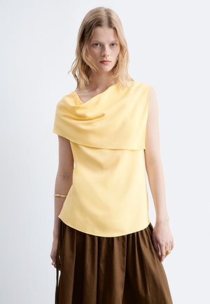 Jeune femme aux cheveux clairs porte un haut jaune sans manches avec un col drapé et une jupe marron foncé sur un fond uni.
