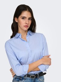 Young woman with long dark hair wearing a light blue button-up shirt and jeans, arms crossed, looking confidently forward.