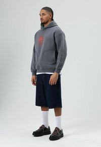 Grijze hoodie met capuchon, een grote voorzak en een rood grafisch ontwerp, gecombineerd met donkere spijkershorts en zwarte schoenen met roze veters.