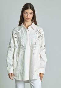 Camisa blanca bordada con diseños florales y de hojas, con cuello y cierre de botones en la parte delantera y mangas largas. Textura de tela suave.